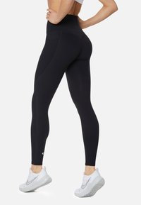 Leggings neri a vita alta con una consistenza liscia, caratterizzati da un design a cucitura e un piccolo logo accanto alla caviglia, abbinati a scarpe da ginnastica bianche.