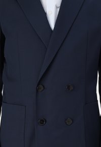 Navy double-breasted blazer in smooth fabric with two front pockets, black buttons, and peak lapels; worn over a light blue striped shirt.
