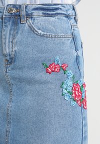 Jupe en denim bleu clair avec broderies florales en rouge, rose et bleu. Dotée d'un design standard à cinq poches et d'une fermeture à bouton en métal.