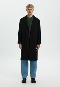 Cappotto di lana nero con colletto a punte, chiusura con bottoni frontali, indossato sopra un maglione verde e jeans a gamba larga azzurri. Scarpe nere.
