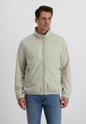 Man met kort donker haar, gekleed in een lichtbeige jas met ritssluiting en blauwe spijkerbroek, staand tegen een effen witte achtergrond.