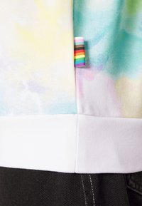 Gros plan sur un tissu tie-dye aux teintes pastel avec une petite étiquette du drapeau arc-en-ciel cousue dans la couture au-dessus d’un ourlet blanc.