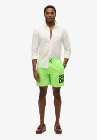 Costumi da bagno verde neon con logo nero "SUPERDRY", cintura elastica e tasche laterali, abbinati a una camicia bianca a maniche lunghe con bottoni.