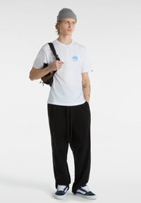 Vit bomullströja med blå Vans-logga, svarta kordfläckbyxor, grå stickad mössa och marinblå sneakers med vita detaljer, hållande en brun väska.