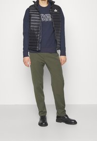 Černý péřový vestový kabát přes tmavomodrou mikinu s bílým grafickým potiskem, olivově zelené kalhoty a černé boty. Hladké textury a přiléhavý design.