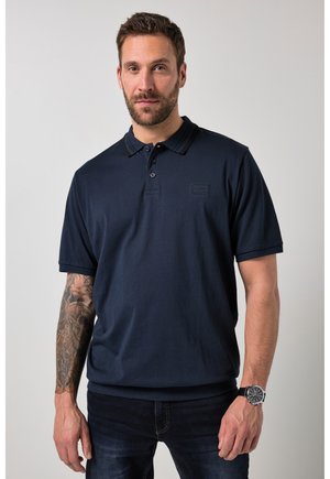 Mand iført navyblå polo, mørke jeans, armbåndsur, synlig detaljeret tatovering på venstre arm, stående mod en ensfarvet baggrund.