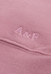 Myk rosa stoff med en strukturert strikk. Har hevet "A&F"-logo i matchende farge, med en ferdig søm nederst.
