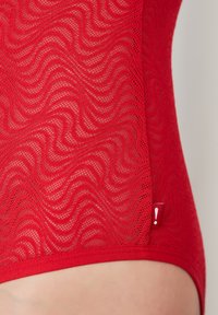 Punainen pitsinen body, jossa aaltoileva kuvio, läpinäkyvä tekstuuri ja sivulla kontrastina pieni punainen etiketti, jossa on huutomerkkki.