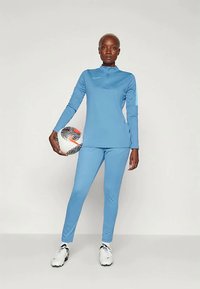 Sportists zilā Nike treniņtērpā tur futbolbumbu vienā rokā, stāvot uz vienkrāsaina baltā fona, valkājot baltas sporta kurpes.