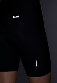 Svarte sykkelshorts laget av glatt stoff, med et hvitt logomerke, tett passform og en liten sidesplitt for pusteevne.