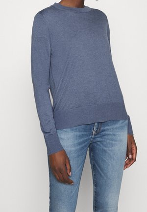 Person iført en blå langærmet rundhals trøje kombineret med lyseblå denimjeans, stående mod en ensfarvet hvid baggrund.