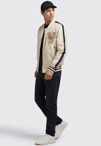 Creme satin bomberjakke med sorte striber på siden, broderet tigerlappen, ribbede manchetter, parret med mørke jeans og hvide sneakers.