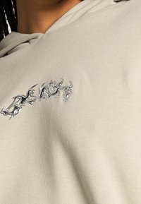 Hoodie beige clair avec un logo métallique argenté sur la poitrine. Le tissu semble doux et lisse avec une coupe décontractée.