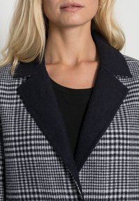 Donna bionda che indossa un top nero e un cappotto a scacchi grigio e nero con ampio colletto scuro, labbra chiuse, sfondo neutro.