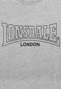 Pelēka kreklu svītra ar lielu melnu "LONSDALE" logotipu, kas izliekts virs "LONDON", drukāts uz mīksta teksturēta auduma.