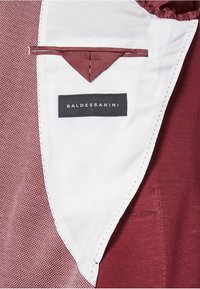 Interno della giacca Baldessarini caratterizzato da un tessuto bordeaux profondo, foderato in bianco, con dettagli cuciti e una piccola tasca con etichetta logo.