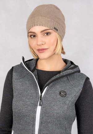 Beige gebreide muts met geribbelde textuur. Model draagt een grijze gezipte bodywarmer over een zwart shirt met lange mouwen, met een logopatch op de bodywarmer.