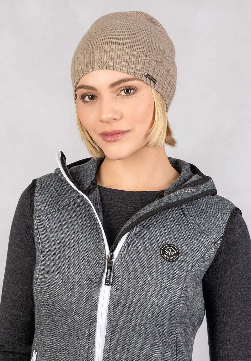 Beige gebreide muts met geribbelde textuur. Model draagt een grijze gezipte bodywarmer over een zwart shirt met lange mouwen, met een logopatch op de bodywarmer.