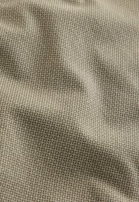 Beige szövet, amelyen kis, ismétlődő geometrikus minta található sötét barna színben. A textúra simának tűnik, finom ráncokkal és redőkkel.