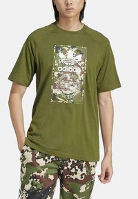 Camiseta de manga corta color verde oliva hecha de tela suave, con un panel de estampado de camuflaje que presenta el logo de Adidas en blanco y detalles en texto.
