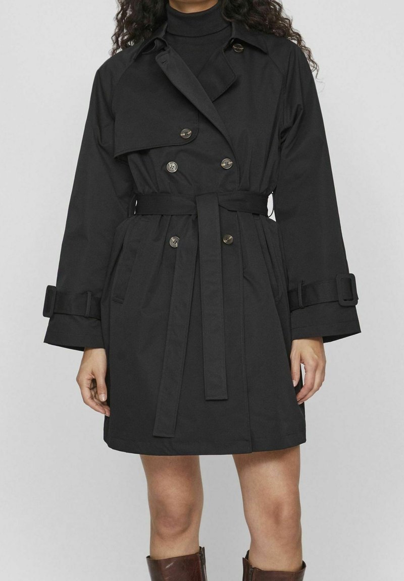 Femme portant un trench-coat noir croisé avec ceinture, poignets boutonnés et col montant, par-dessus un col roulé noir, assorti à des bottines marron.