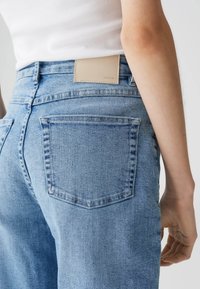 Jeans in denim azzurro chiaro con vita alta, dotati di una linguetta rettangolare in pelle beige e tasche posteriori. Texture liscia e cuciture classiche.