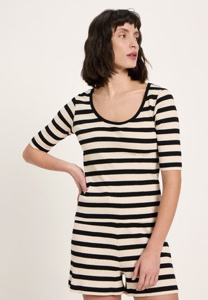 Frau mit schulterlangem dunklem Haar, die einen kurzärmligen Einteiler mit schwarzen und weißen horizontalen Streifen trägt, schaut zur Seite und hat eine Hand in die Hüfte gestützt.