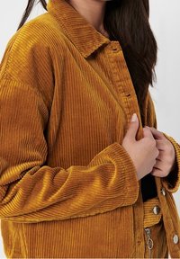 Giacca in corduroy senape con texture a costine, dotata di colletto, chiusura a bottoni e tasche laterali.