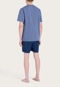 Camicia a maniche corte a quadri blu e bianchi abbinata a pantaloni corti blu navy, entrambi realizzati in tessuto leggero. Piedi nudi.