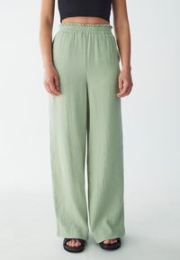 Pantalones de pernera ancha verde claro, confeccionados en una tela texturizada y con cintura elástica. Llevado con sandalias planas negras, con un diseño de punta abierta.