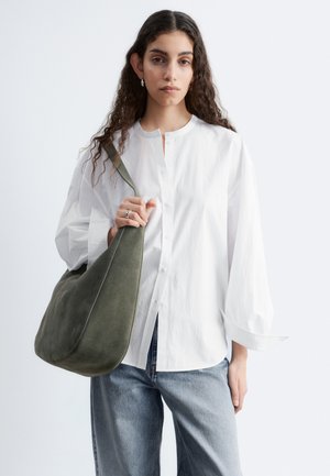 Femme marchant portant une chemise blanche ample boutonnée, un jean bleu clair, des sandales noires et portant un grand sac à bandoulière vert olive.