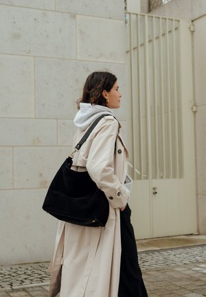 Woman wearing beige trench coat and gray hoodie, carrying large black shoulder bag, walking past a light stone wall and metal gate.
