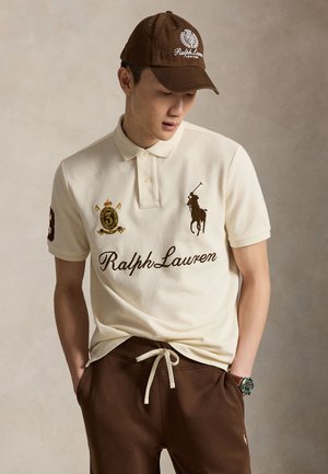 Homme portant un polo blanc Ralph Lauren avec des logos, une casquette marron et un pantalon marron à cordon, regardant vers le bas, les mains dans les poches.