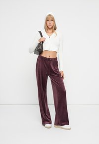Leichter, kurzer weißer Pullover mit Kapuze, kombiniert mit lila Samt-Hose mit weitem Bein und elastischem Bund. Getragen mit weißen Schuhen.