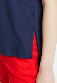 Granatowa bluzka z krótkim rękawem i bocznymi rozcięciami, w zestawieniu z czerwonymi spodniami. Materiał wydaje się gładki i lekki, o swobodnym kroju.