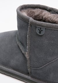 Bottine grise en daim avec une doublure en fourrure douce, présentant un design de coutures cousues et un accent logo noir près du col. Texture lisse.