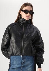 Veste bomber en cuir noir avec un col haut, fermeture éclair et coupe oversize. Associée à une jupe en denim taille haute.