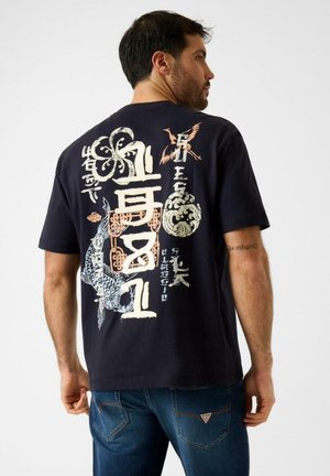 Hombre con camiseta negra con diseños gráficos asiáticos en blanco y naranja y pantalones vaqueros azules, de espaldas contra un fondo liso.