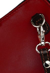 Hoek van een rode leren handtas met een zilverkleurige sluithaak, voorzien van een bruine band met stikseldetail. Glad oppervlak.