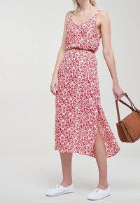 Robe maxi à imprimé floral rouge avec des bretelles spaghetti, une taille marquée, une fente sur le côté et un tissu léger, accompagnée d'un sac marron et de chaussures blanches.