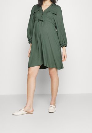 Kobieta ubrana w luźną, sięgającą kolan oliwkowo-zieloną sukienkę i kremowe wsuwane mokasyny, stojąca na jednolitym białym tle.