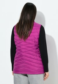 Lila gesteppte Weste mit Chevron-Muster, aus leichtem Material gefertigt, kombiniert mit einem schwarzen Langarmoberteil. Sanfte Textur und entspannte Passform.