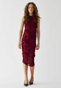 Burgundy ärmlös klänning med blommönsterbroderi, rynkad tyg och knälång design. Bärs med svarta högklackade skor.