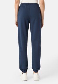 Tmavě modré tepláky vyrobené z měkkého materiálu, s elastickým pasem a vypnutými kotníky. Nosené s bílými teniskami.