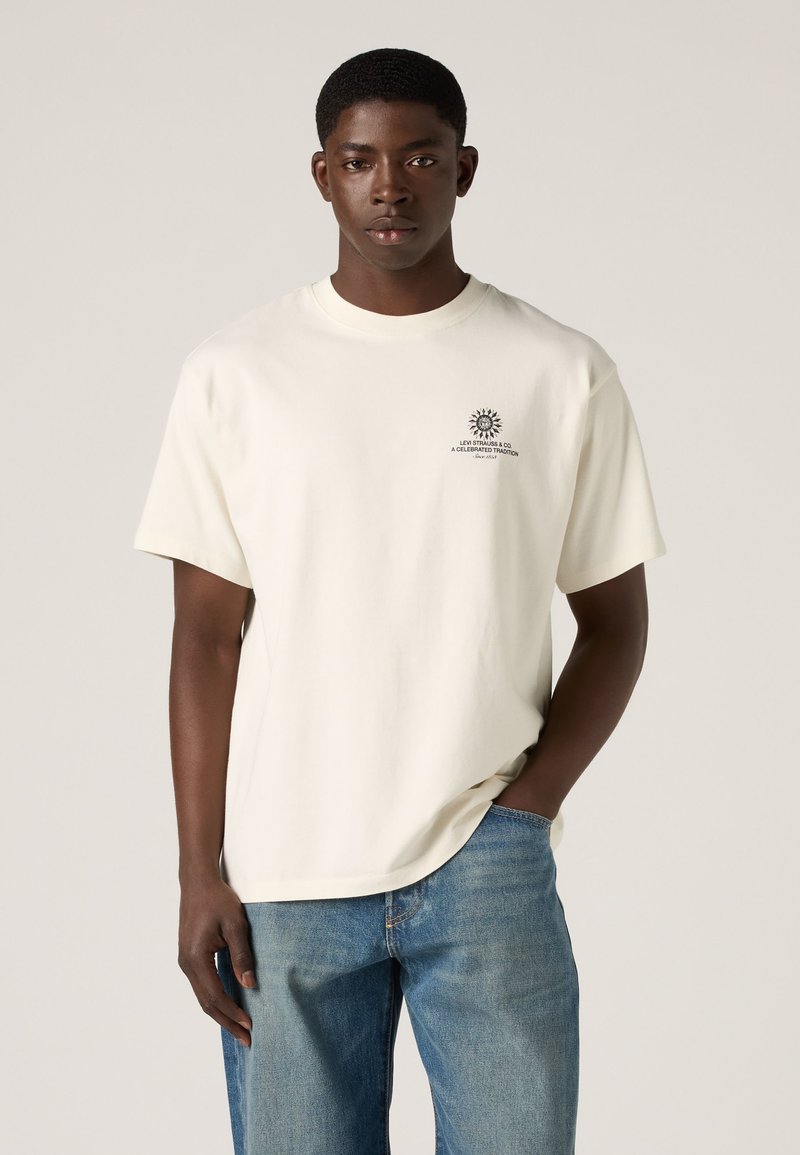 Young man wearing a white short-sleeve t-shirt with small chest print and blue jeans, standing with one hand in pocket.