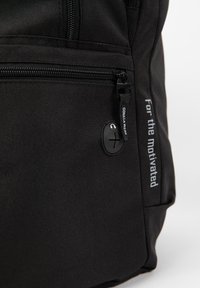 Zwarte stoffen rugzak met een rits, een rond logo en de tekst "For the motivated" in het wit gedrukt op de zijkant.