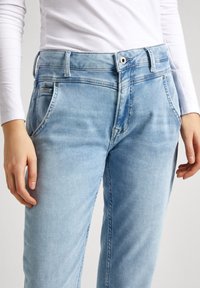 Los jeans de mezclilla azul claro presentan un diseño clásico de cinco bolsillos, pasadores para el cinturón y un cierre de cremallera con botón. Textura suave y corte relajado.