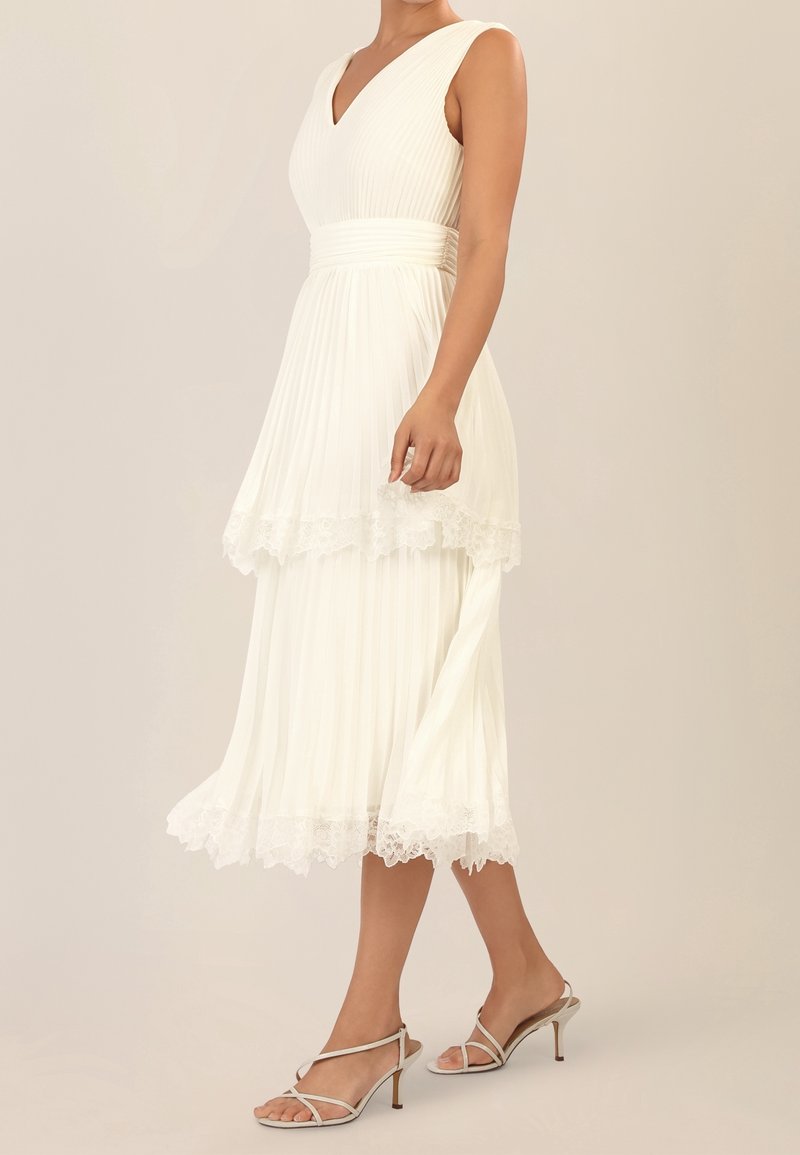 Femme portant une robe midi blanche plissée sans manches avec une bordure en dentelle et des sandales beiges à talons hauts sur un fond uni.