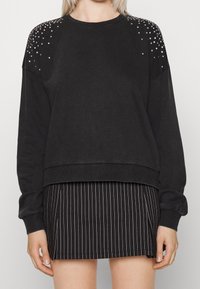 Sudadera negra con detalles de tachuelas en los hombros, cuello redondo y puños acanalados. Combinada con una falda a rayas en blanco y negro.