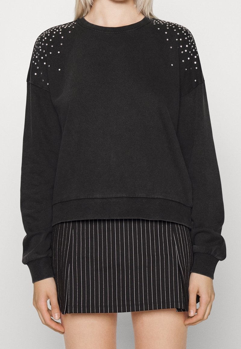 Sudadera negra con detalles de tachuelas en los hombros, cuello redondo y puños acanalados. Combinada con una falda a rayas en blanco y negro.